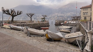 Un muelle con barcos amarrados, un paseo de piedra y vista a un pueblo y montañas a lo lejos.