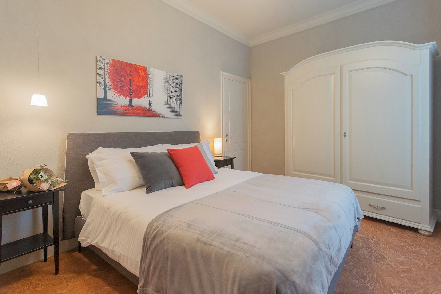 A bedroom with a large bed, a nightstand with a lamp, a painting on the wall, and a wardrobe.