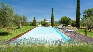 Une piscine rectangulaire avec une terrasse en bois, entourée de champs de lavande, et dotée de chaises longues et d’une table avec des parasols.