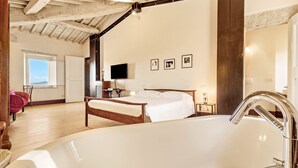 Une chambre spacieuse avec une grande baignoire blanche, des poutres en bois au plafond, un lit, un bureau et un téléviseur fixé au mur.