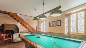 Une table de billard dans une pièce dotée d’une cheminée, de poutres en bois et de grandes fenêtres.