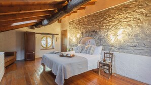 Une chambre avec un mur en pierre, des poutres en bois au plafond, un lit avec des oreillers, une table de chevet et une fenêtre.
