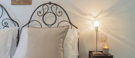 Une chambre à coucher avec un lit en métal, du linge de lit blanc, une table de chevet avec une lampe et un luminaire fixé au mur.