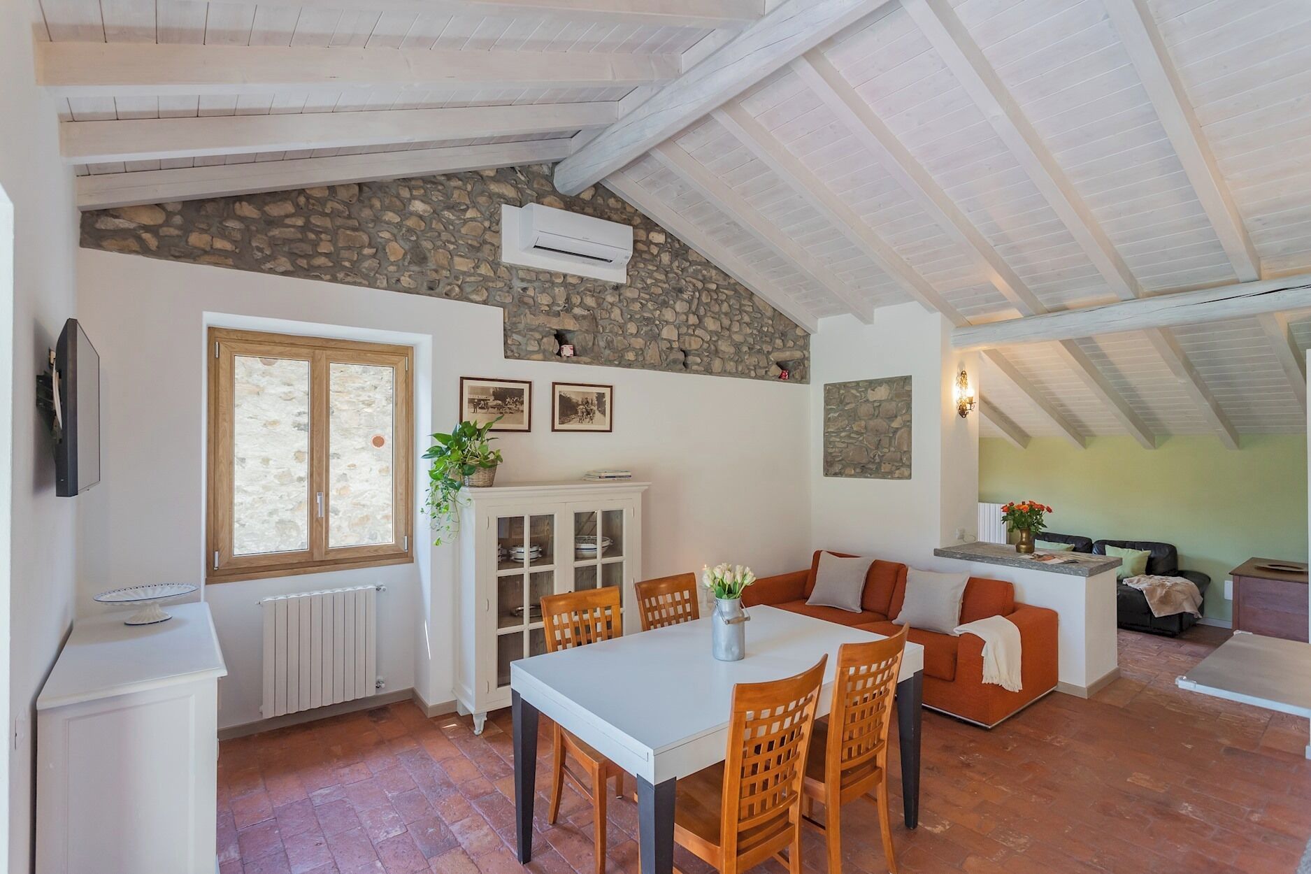 Un intérieur rustique avec un mur en pierre, des poutres en bois, un coin repas avec une table et des chaises, un canapé et un téléviseur fixé au mur.