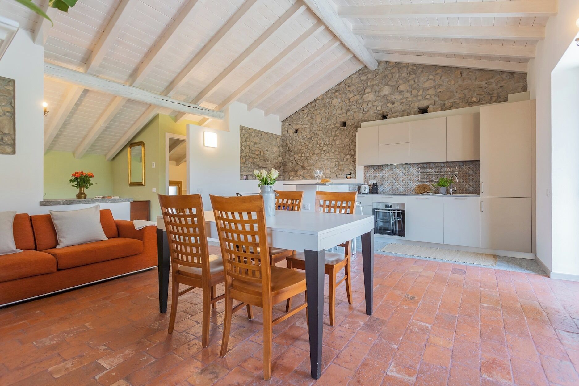 A dining area with a table and chairs, a kitchen with white cabinets, and a stone wall.