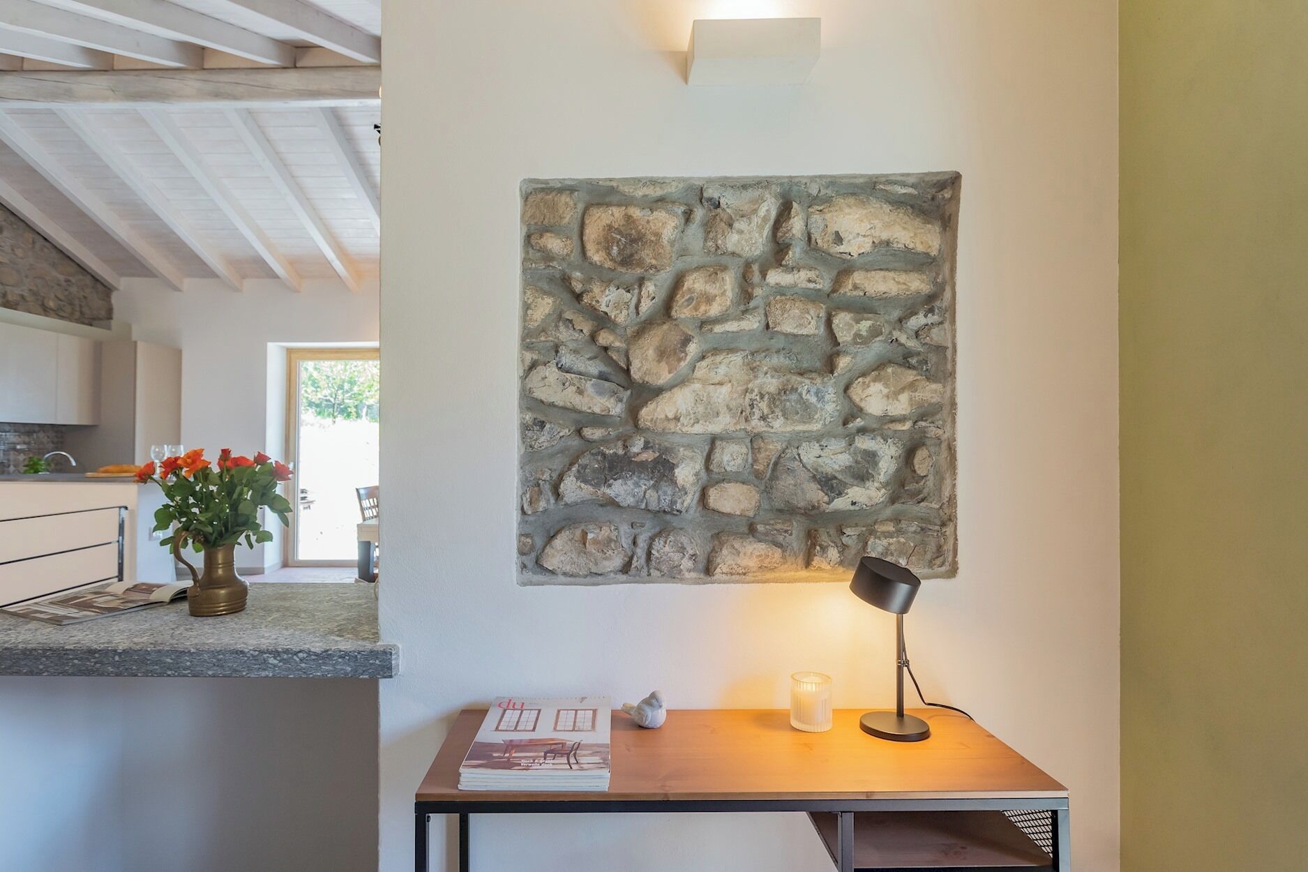A modern kitchen with a stone wall art piece, a wooden table with a lamp, and a vase with flowers.