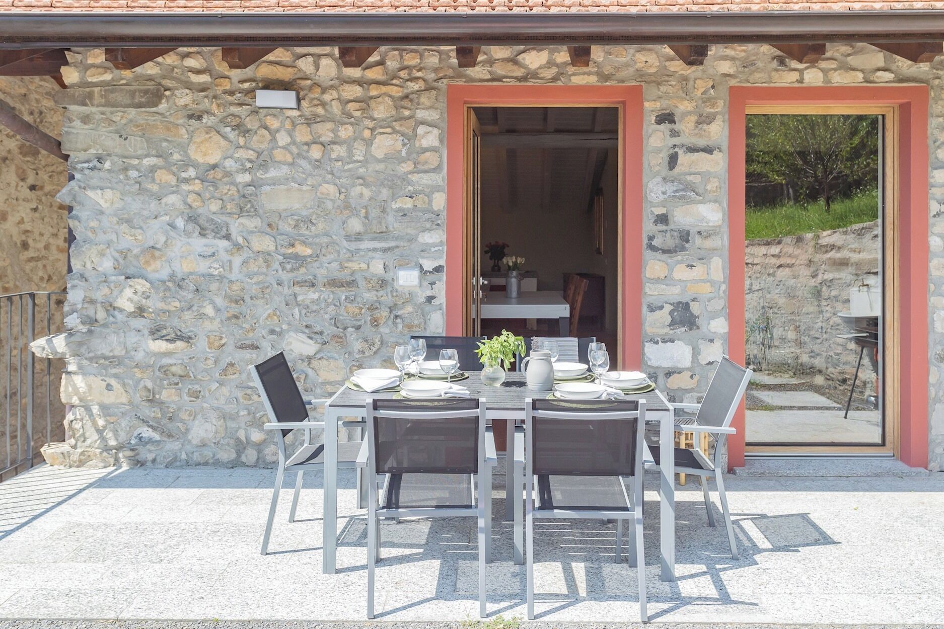 Un espacio de comedor al aire libre con un muro de piedra, una mesa lista para cuatro comensales y dos puertas que dan al interior.