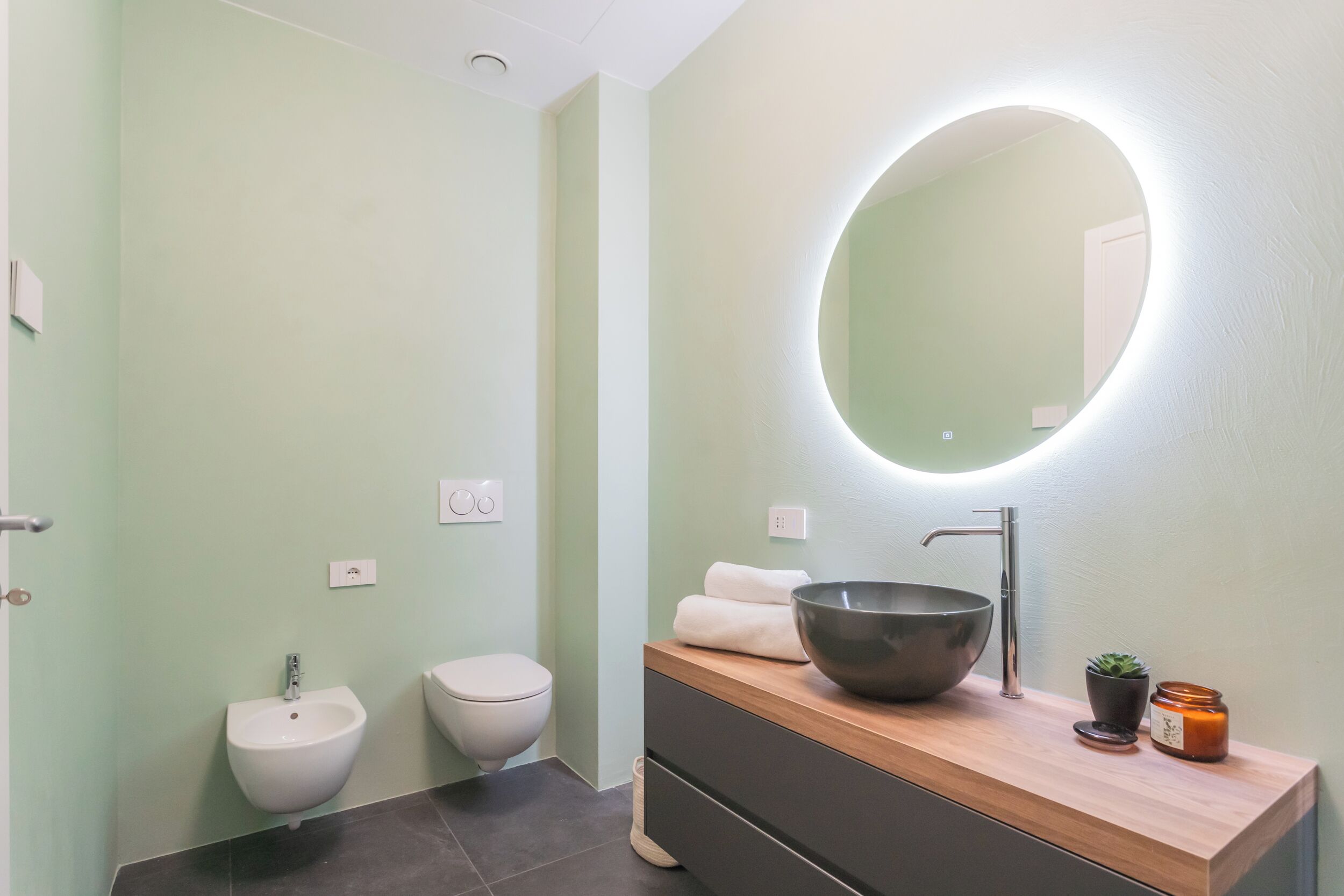 Une salle de bain moderne avec un grand miroir rond, un bidet, des toilettes et un meuble-lavabo en bois doté d’un vasque.