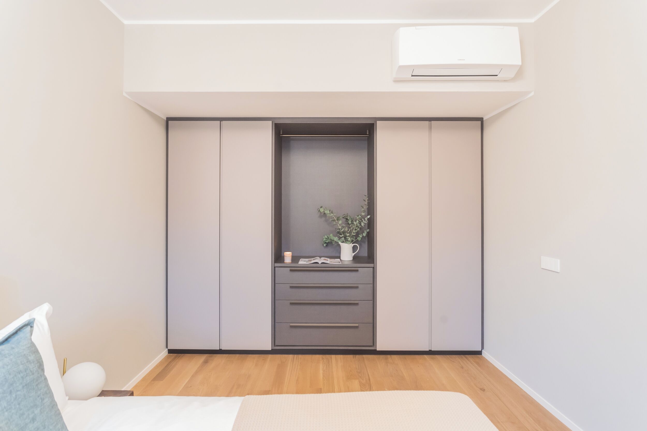 A modern bedroom with a built-in wardrobe, a bedside table with a lamp, and a small plant on the dresser.