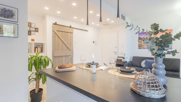A modern kitchen with a dark countertop, a wooden door, and a dining area with a grey sofa and a vase of greenery. - We Lake Como by Rent all Como (Argegno)