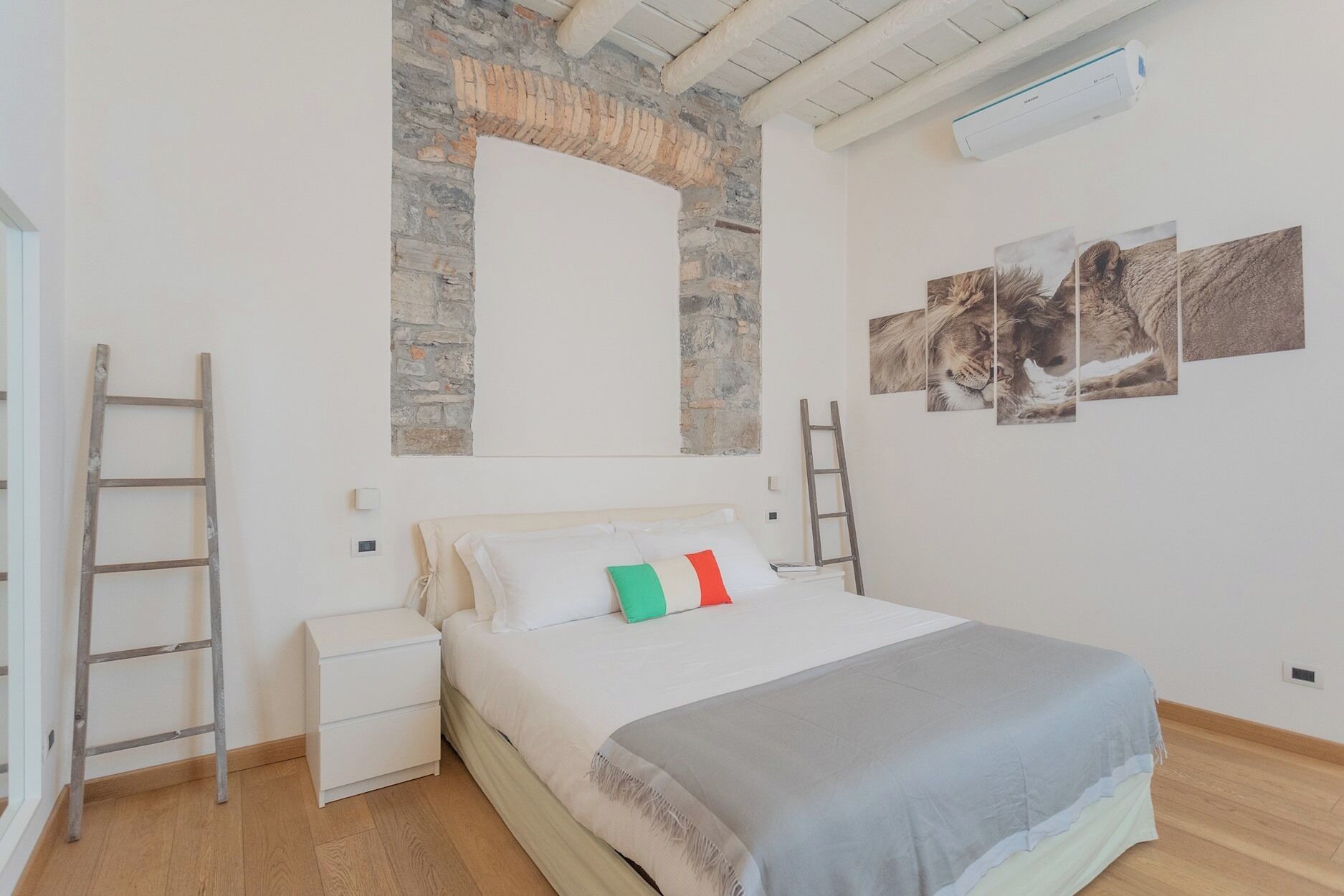 A bedroom with a stone wall, a bed with a grey blanket, and three pillows. There are two ladders against the wall and a white bedside table.