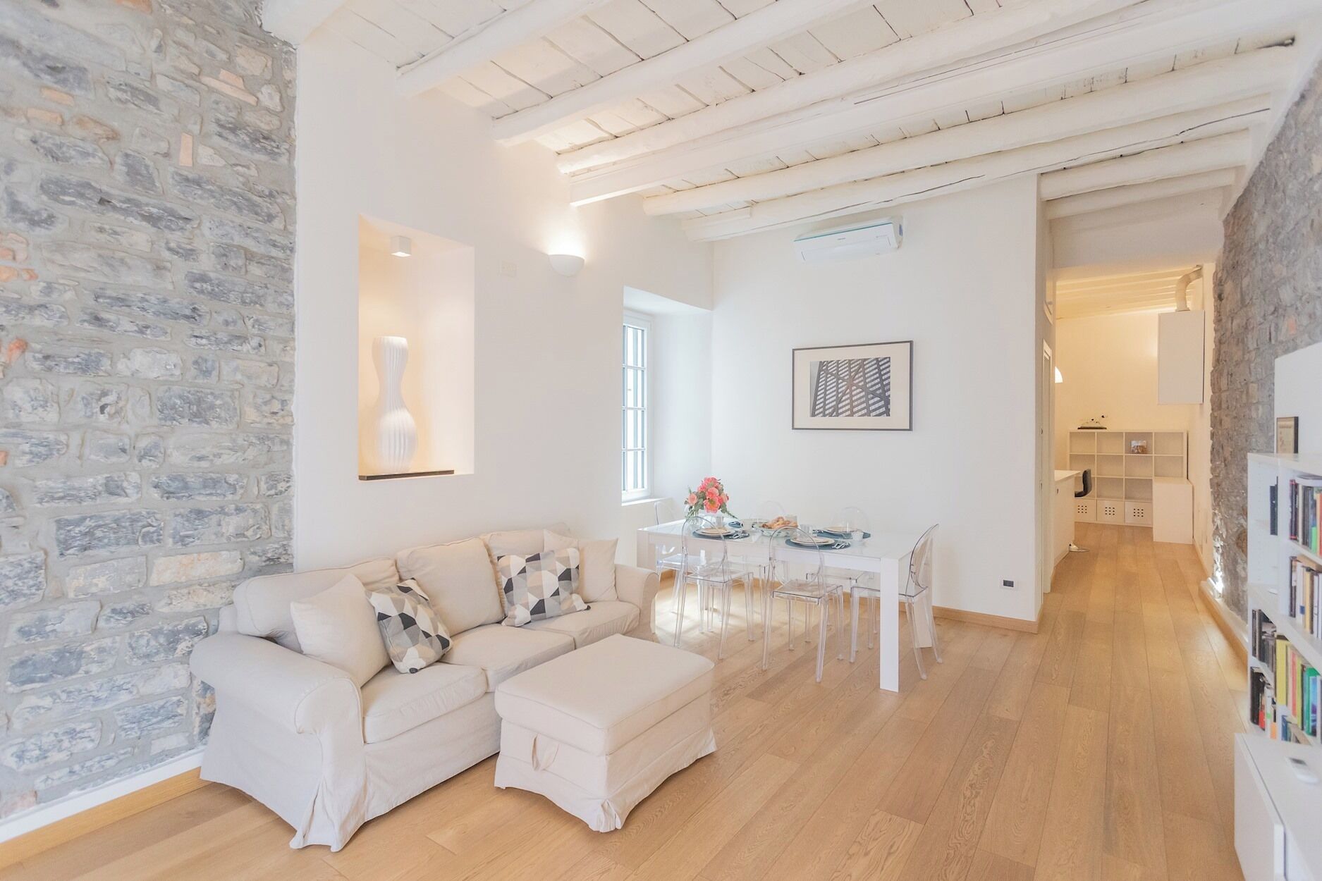 A spacious living area with a stone wall, a white sofa set, a dining table with chairs, and a bookshelf.
