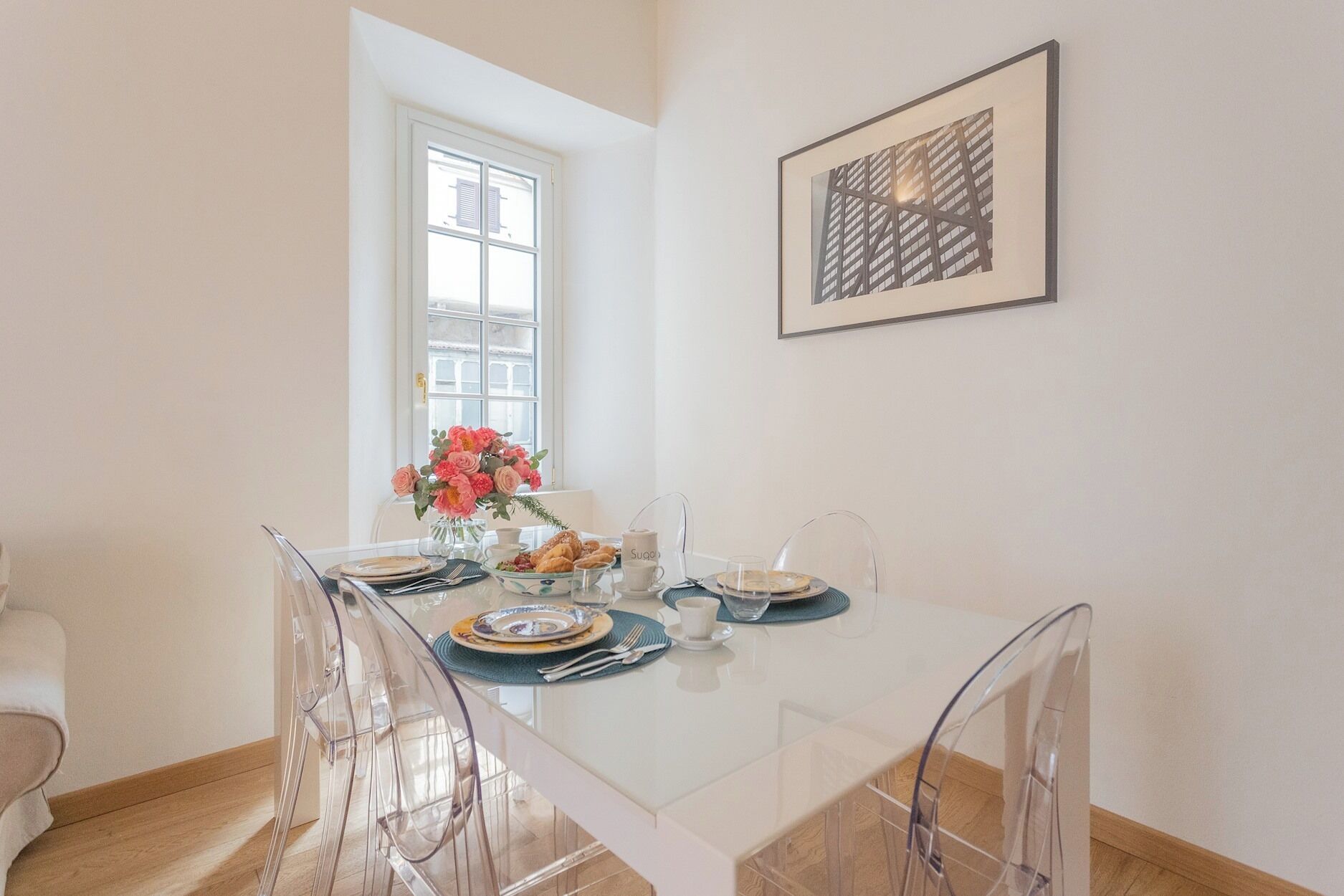 Ein Essbereich mit Glastisch, transparenten Stühlen, einer Blumenvase und einem gerahmten Bild an der Wand.