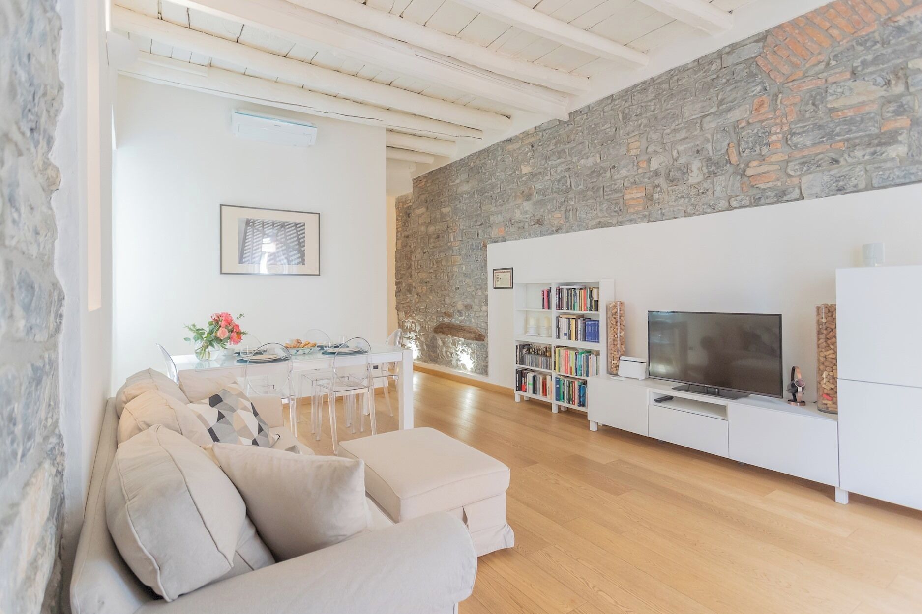 Una sala de estar moderna con pared de piedra, un sofá blanco, una zona de comedor con mesa y sillas, y una estantería con un televisor.