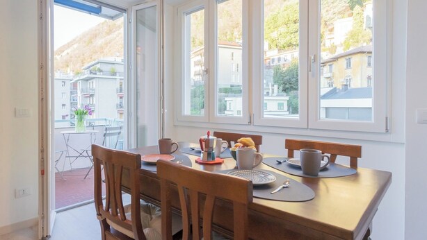 A dining area with a table set for two, large windows with a view of buildings and trees, and a door leading to an outdoor balcony. - White Lake View Apartment by Rent all Como (Como)