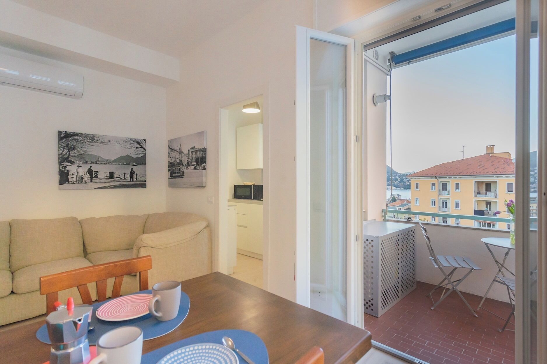 Un salon confortable avec un canapé, une table à manger et des chaises. Un balcon avec vue sur les bâtiments et le paysage urbain est visible à travers une porte ouverte.