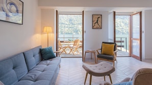 A modern living room with a sofa, a small table, and a chair. There is a large window with a view of a beach and trees. - Your Private Harbour Nearby George Clooney by Rent all Como (Carate Urio)