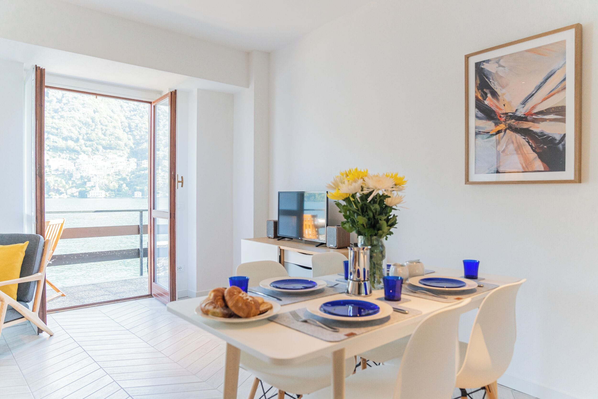 Un moderno comedor con una mesa preparada para dos, un jarrón con flores amarillas y vistas al exterior a través de un ventanal.