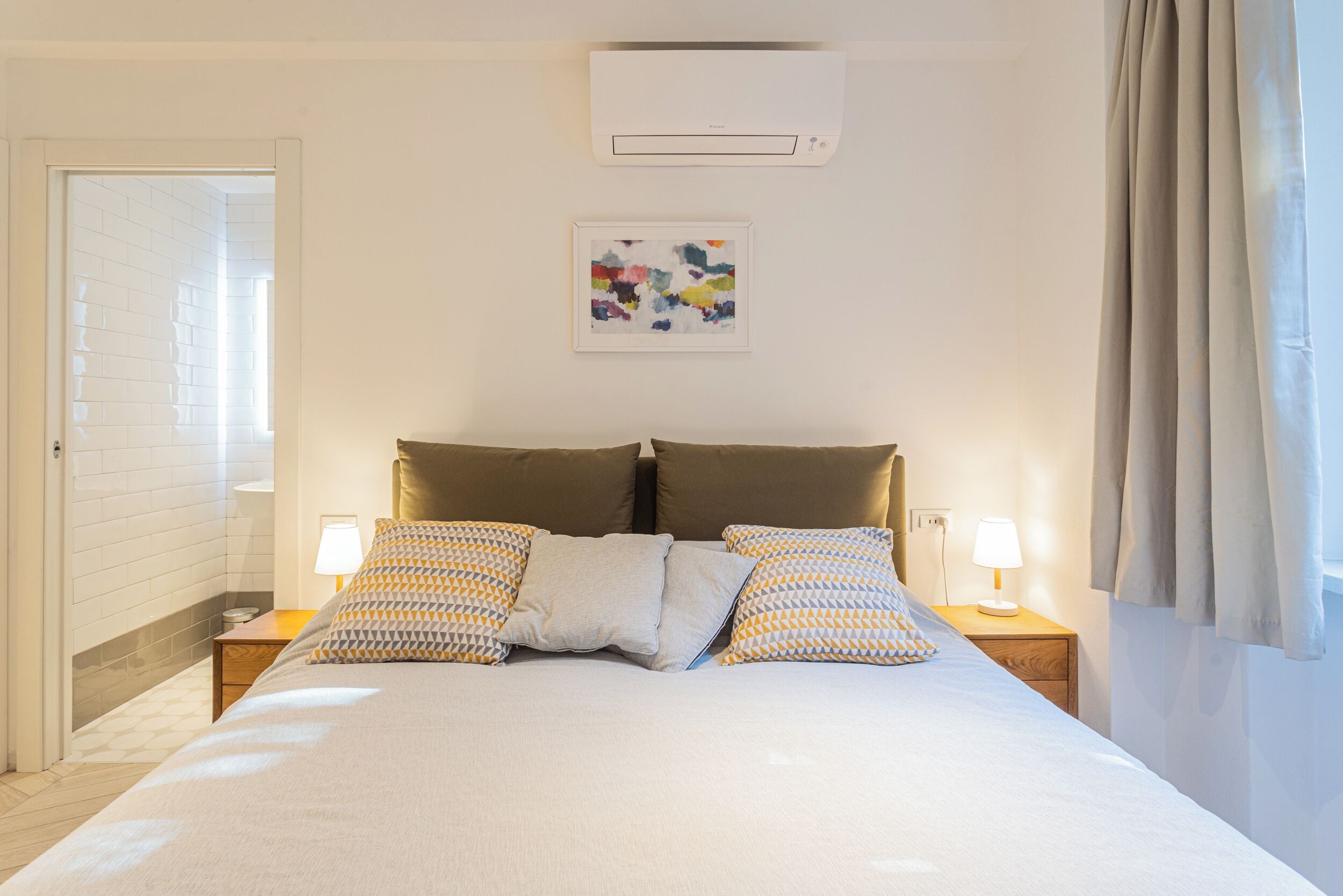 A neatly arranged bedroom with a bed, two bedside tables, a wall-mounted air conditioner, and a framed abstract painting.