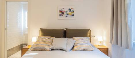 A neatly arranged bedroom with a bed, two bedside tables, a wall-mounted air conditioner, and a framed abstract painting.