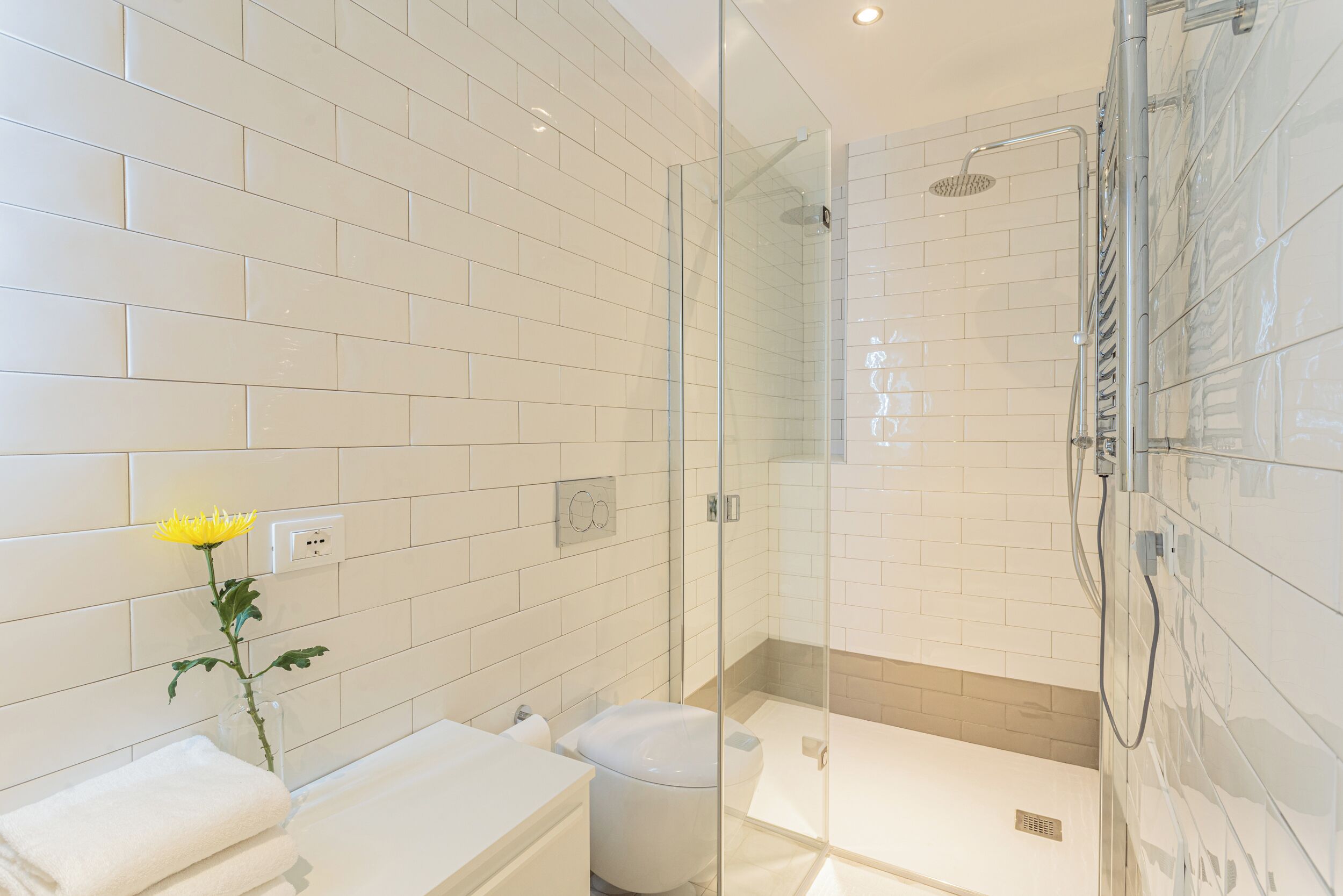 A modern bathroom with white tiles, a glass shower enclosure, a white toilet, and a vase with a yellow flower.