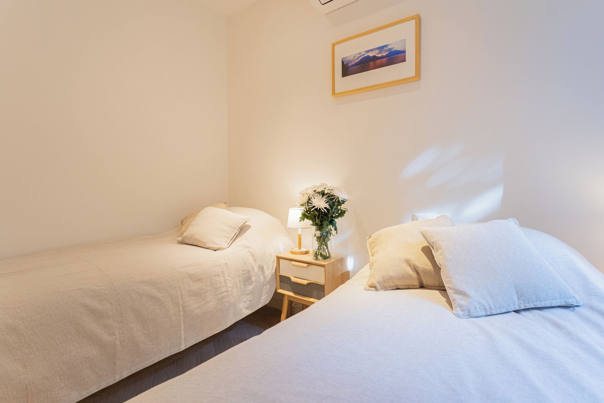 Une chambre avec deux lits simples, une table de chevet avec une lampe et des fleurs, et un tableau encadré au mur.