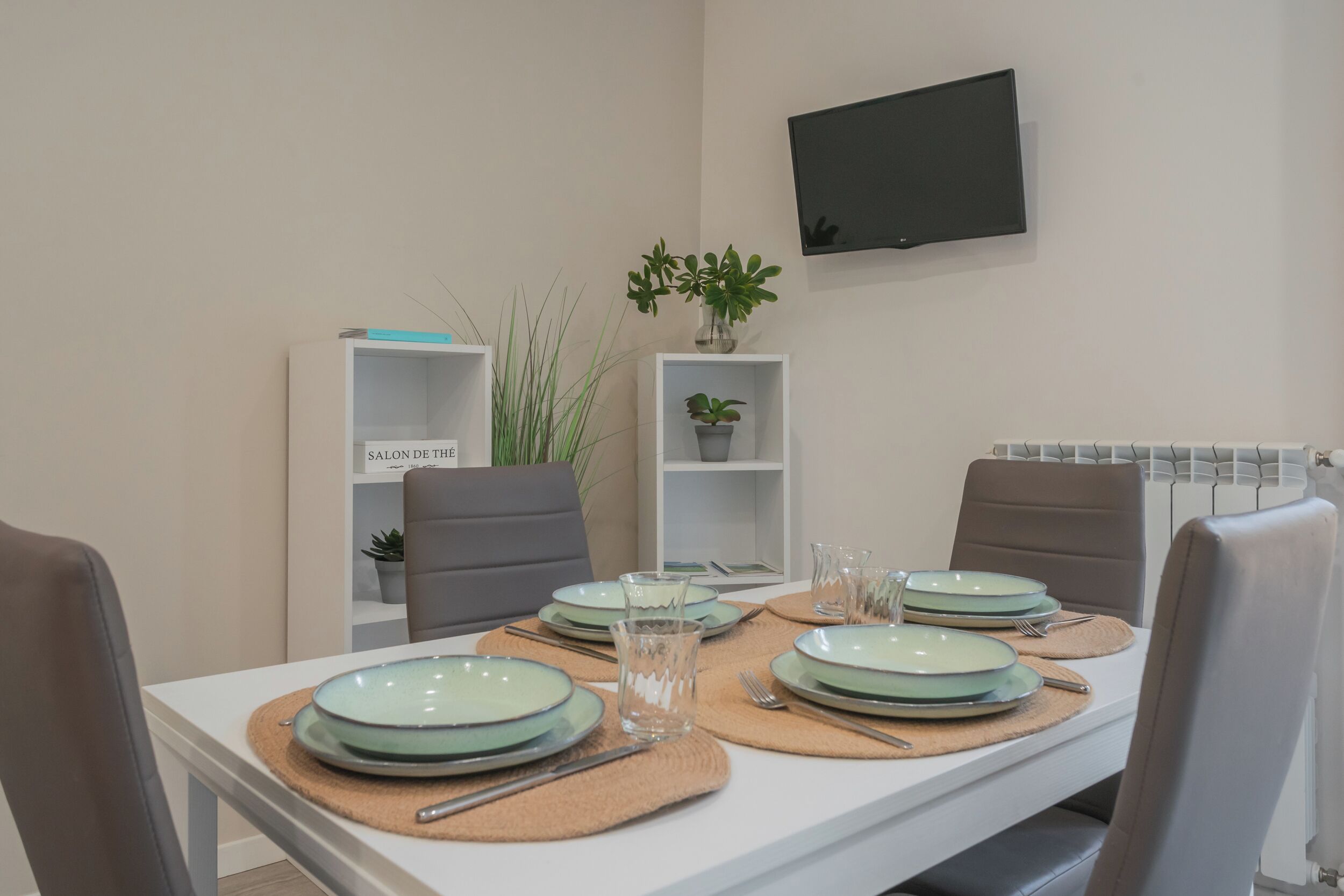 A dining table set with plates, cutlery, and glassware, flanked by chairs, with a TV mounted on the wall and shelves with plants in the background.