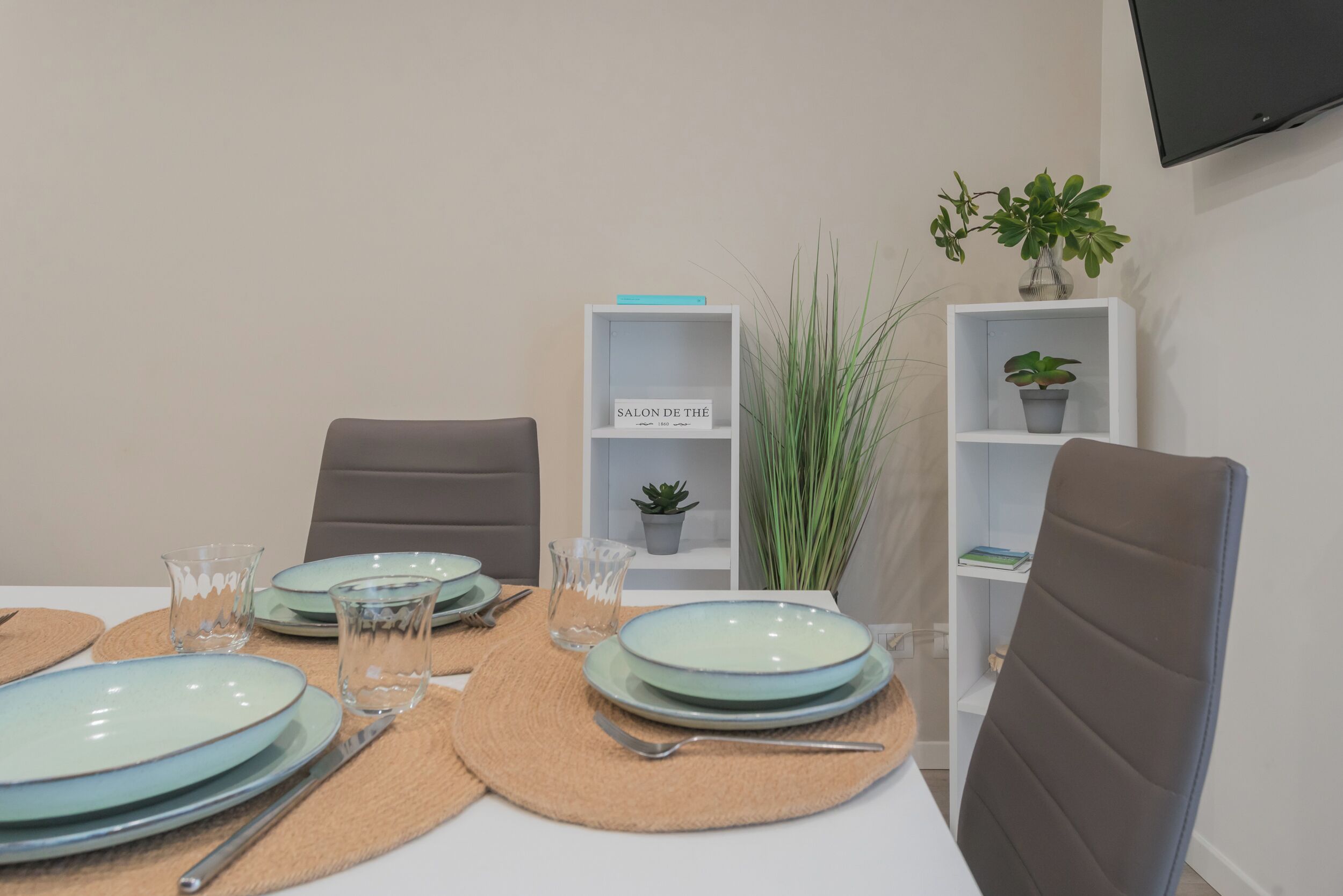 A dining table set with plates, cutlery, and glassware, flanked by chairs and shelves with plants.