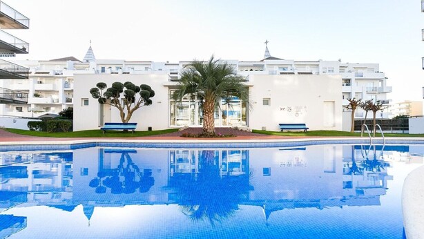 A swimming pool with clear blue water, surrounded by white buildings and palm trees. - BLAUMARINA 225 Roses (Rosas / Roses)