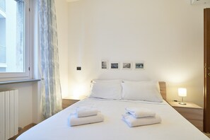 A hotel room with a bed, two white towels, a nightstand with a lamp, and a window with curtains.
