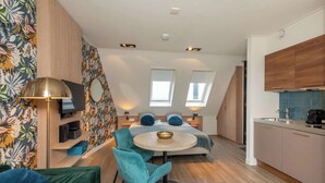 Ein kompakter, moderner Raum mit einem Bett, einem kleinen runden Tisch, zwei Stühlen, einem Fernseher und einer Küchenzeile.