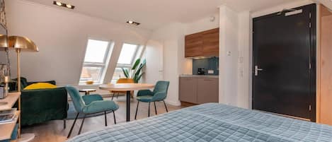 A compact hotel room with a bed, a dining table, two chairs, a small kitchen area, and a large window providing natural light.