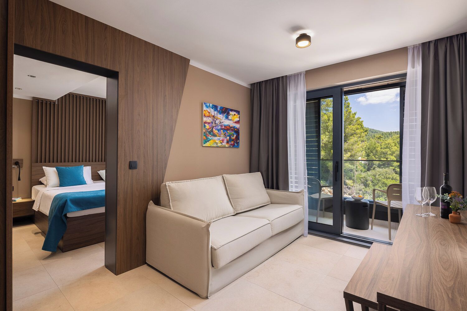 A modern hotel room with a bed, a sofa, a dining table, and a balcony with a view.