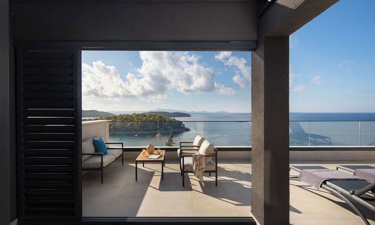 A balcony with a table, chairs, and a view of the sea and hills.