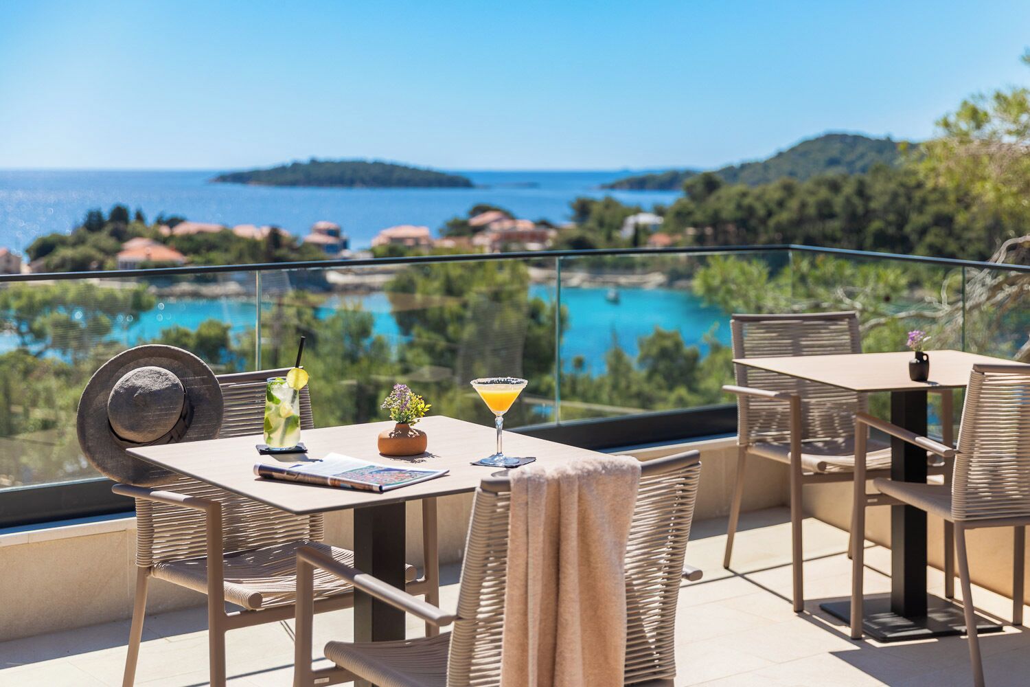 A balcony with a table and chairs, a drink, and a view of a body of water and greenery.