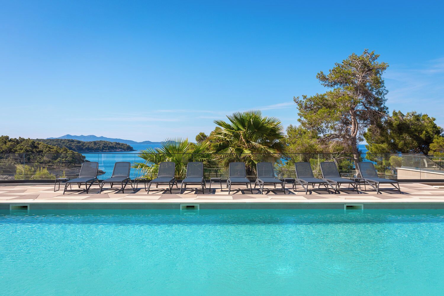 A pool with clear blue water, surrounded by lounge chairs and palm trees, with a view of the sea and hills in the background.