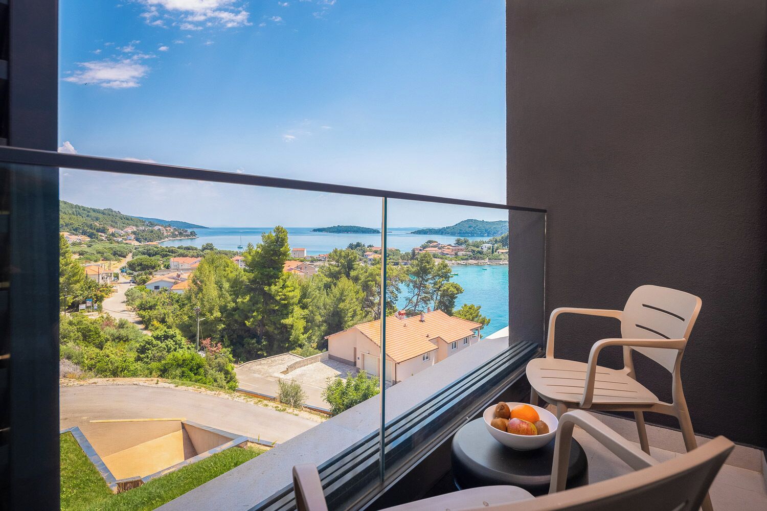 A balcony with a clear view of a coastal town, a bowl of fruit on a table, and two chairs.