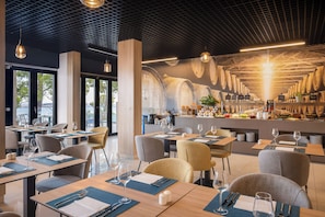 A restaurant with a modern interior, featuring tables with blue placemats, chairs, and hanging pendant lights. - Aminess Alfir Hotel (Blato)