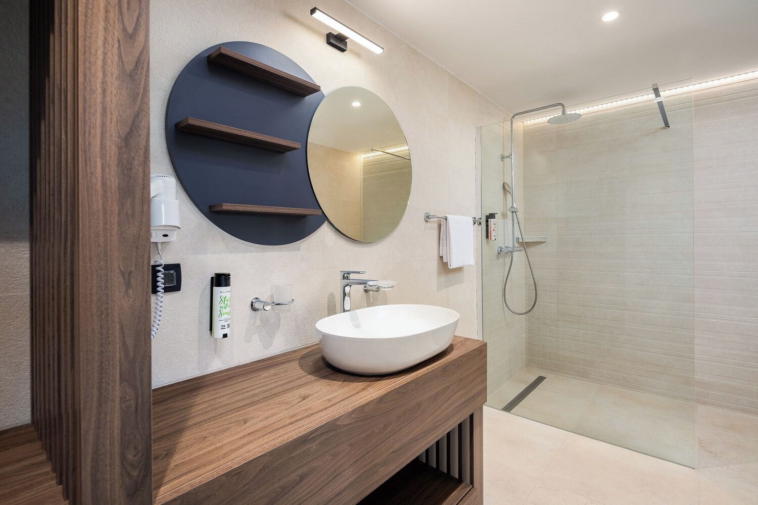 A modern bathroom with a round mirror, a wooden vanity, a white sink, and a glass shower enclosure.