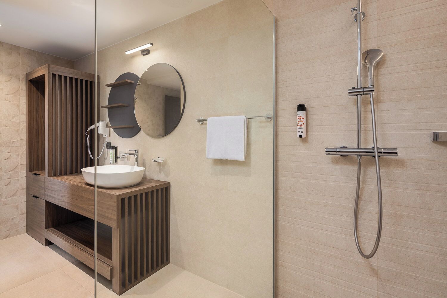 A modern bathroom with a wooden vanity, a round mirror, a white sink, and a glass-enclosed shower area.