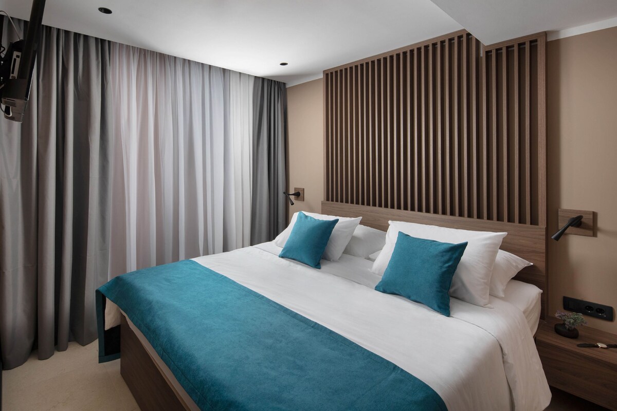 A modern hotel room with a large bed, a wooden headboard, a nightstand, and a wall-mounted lamp.