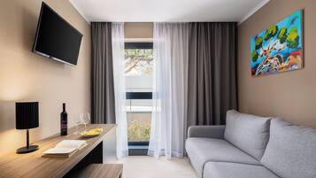 A modern hotel room with a flat-screen TV, a wooden desk with a wine bottle and glasses, a grey sofa, and a large window with sheer curtains.