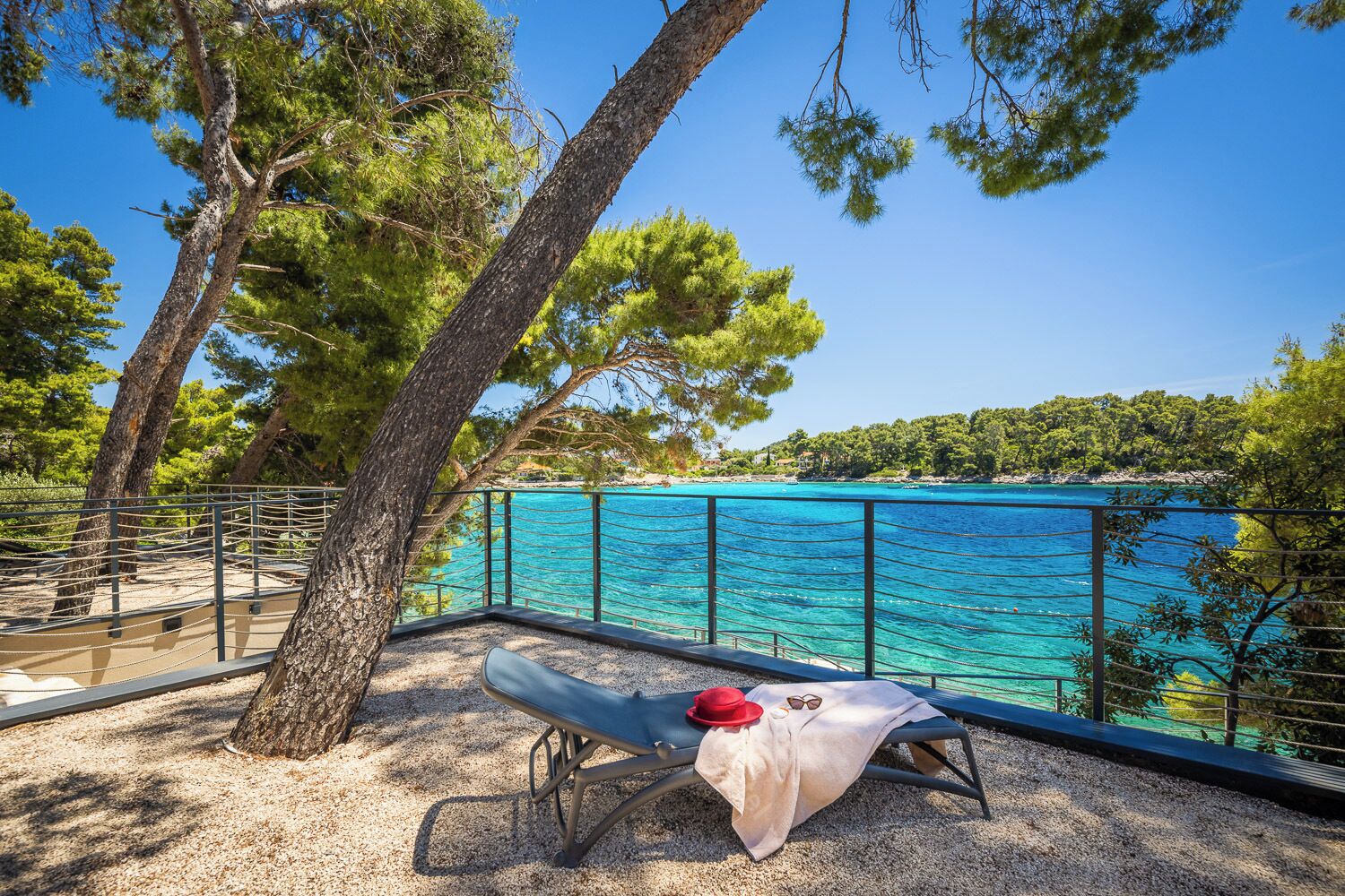 A beachside lounge with a view of a turquoise sea, surrounded by trees and a metal railing.