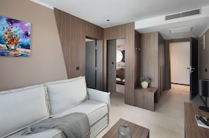 A modern living room with a white sofa, a coffee table, and a painting of flowers. There is a door leading to a bathroom with a mirror and sink. - Aminess Alfir Hotel (Blato)