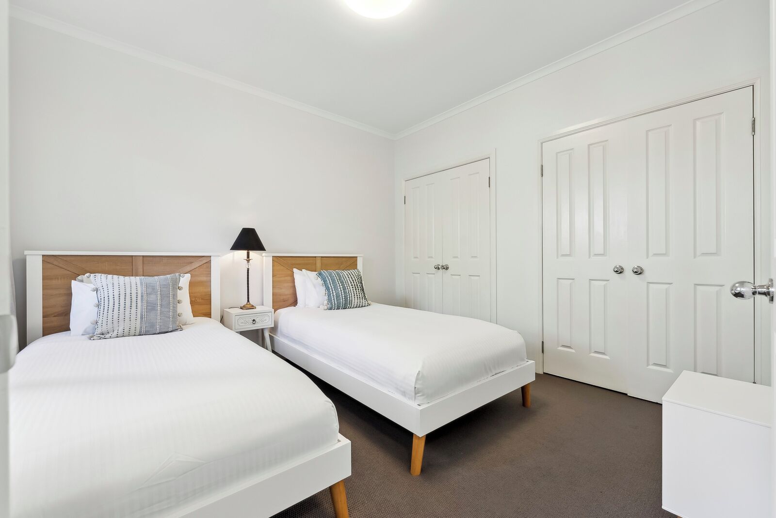 Una habitación de hotel con dos camas individuales, una mesita de noche con lámpara y un armario blanco.