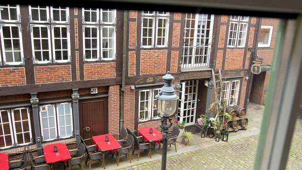 A view of a building with multiple windows and a brick facade. There are tables and chairs outside, some with red tablecloths. A street lamp is visible in the foreground. - Elbphilharmonie Suite (Hamburg)