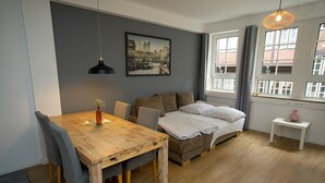 A modern room with a wooden dining table, a sofa bed, a small white side table, and a large window with curtains. - Elbphilharmonie Suite (Hamburg)