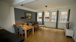 A modern living room with a wooden dining table, grey chairs, a sofa with cushions, a small white side table, and a white cabinet. - Elbphilharmonie Suite (Hamburg)