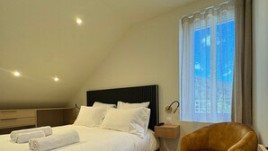 A neatly made bed with white linens and two rolled towels, a wooden nightstand with a lamp, a modern armchair, and a window with curtains. - Residence VillaBelleFleur-Apartment L'Edelweiss (Château-Ville-Vieille)