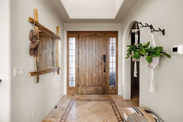 Un couloir avec une porte en bois, un miroir, un support à plantes en macramé et un tapis à motifs.